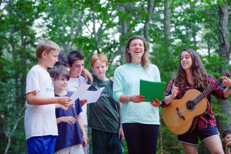 A group of Chewonki staff and campers sing and play music in the woods.